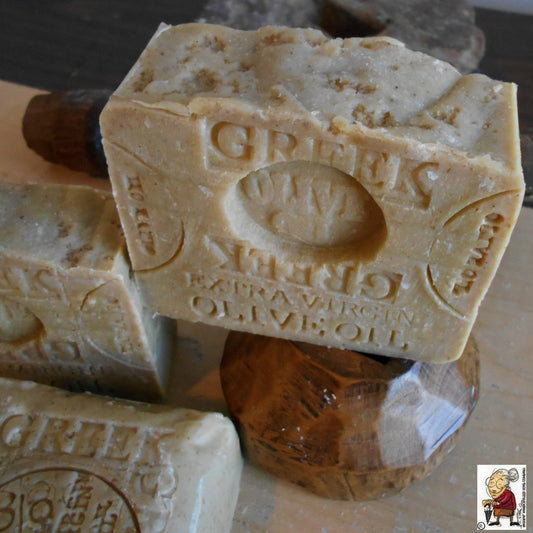Close-up of two bars of Greek extra virgin olive oil soap, enhanced with Mediterranean Sea Salt. One bar is propped up and detailed with an engraved label. The soap bars have a natural, rustic appearance, with some texture on the surface, and are displayed on a wooden stand.