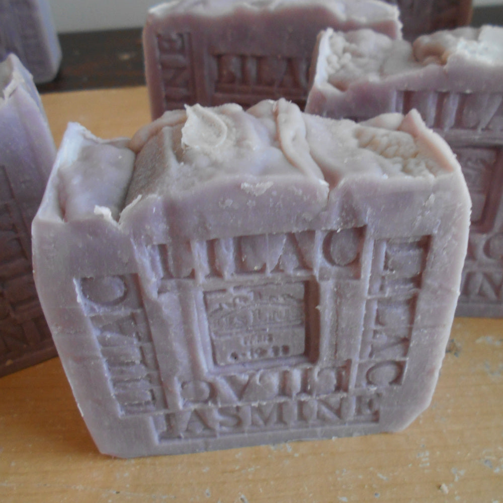 A detailed bar of French Jasmine Lilac Soap with Organic Shea Butter embossed with "Lilac" and "Jasmine" on its surface. The square-shaped bar, featuring a crown motif and the text “NEST.LAIT” and “Paris 1823,” is purple with a textured, rustic appearance. 