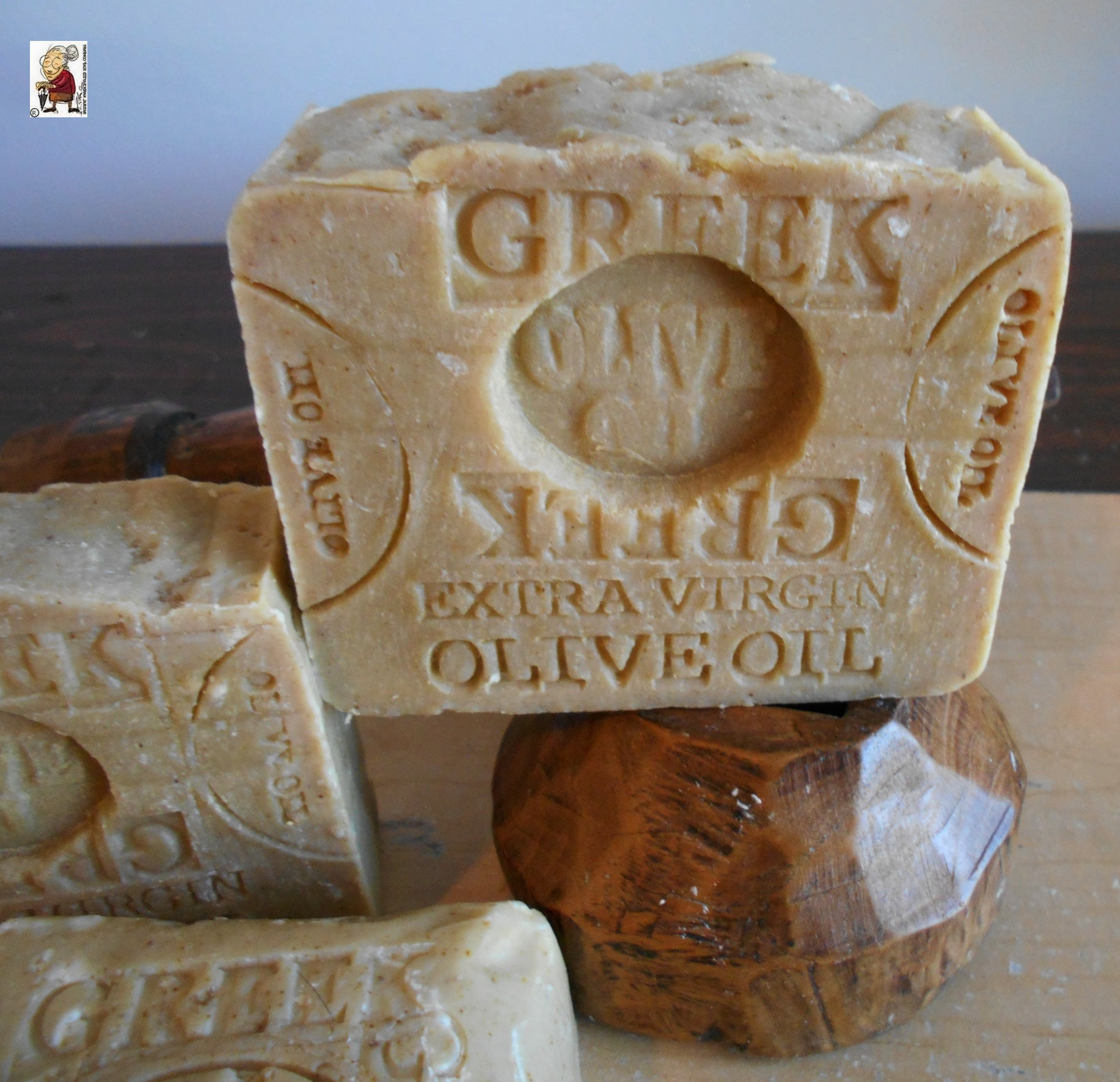 Close-up of two bars of Greek extra virgin olive oil soap, enhanced with Mediterranean Sea Salt. One bar is propped up and detailed with an engraved label. The soap bars have a natural, rustic appearance, with some texture on the surface, and are displayed on a wooden stand.