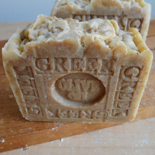 Close-up of two bars of Greek extra virgin organic olive oil soap. One bar is propped up and detailed with an engraved label. The soap bars have a natural, rustic appearance, with some texture on the surface, and are displayed on a wooden stand.